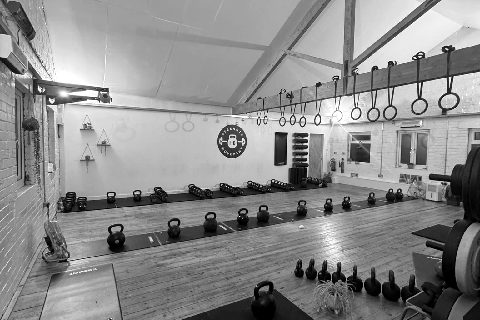 The studio, empty: kettlebells set out on mats along a wooden floor, gymnastic rings hanging from a roof beam, and the Strength & Movement logo on the far wall